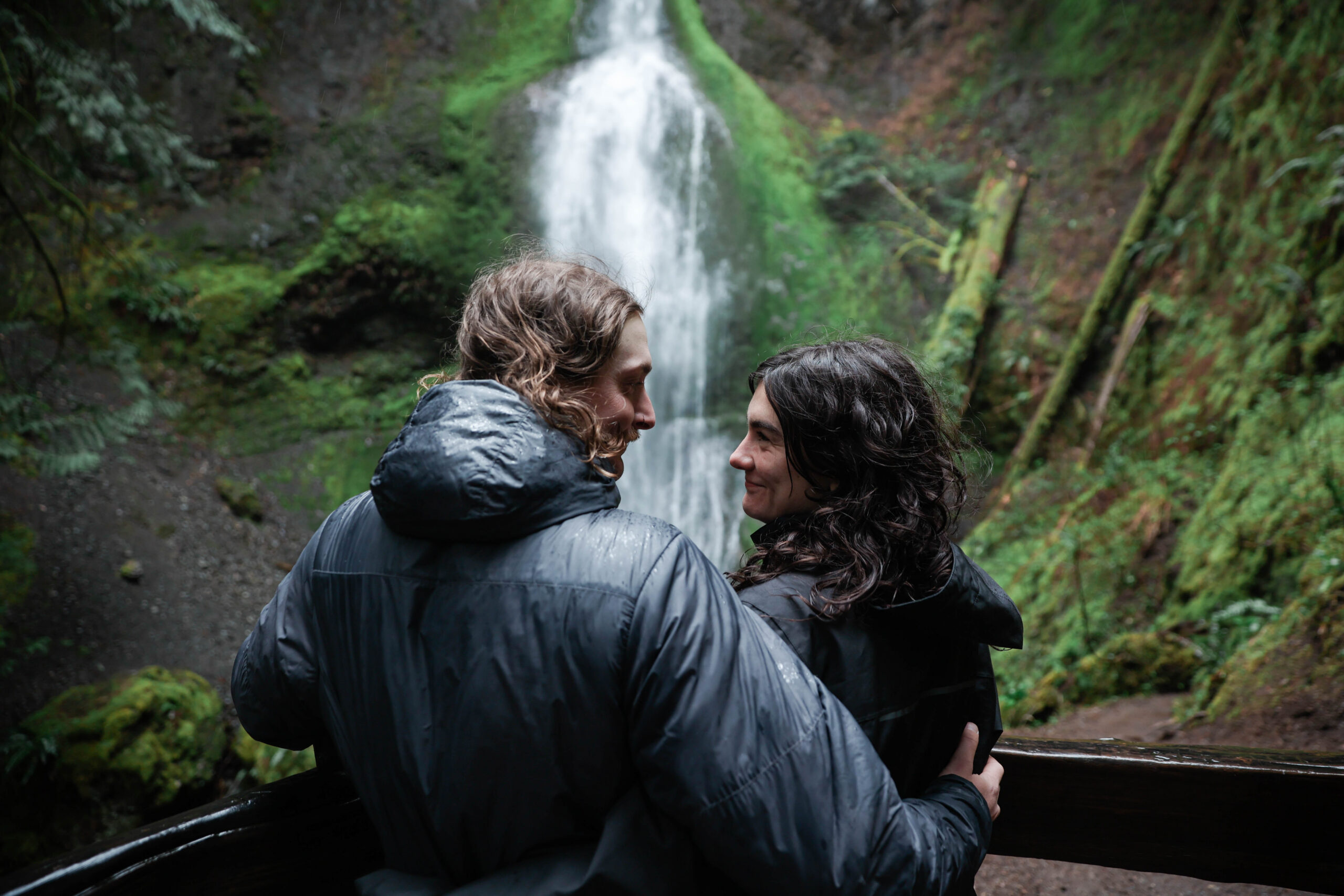 A couple smiles at each other in the rain in front of Marymere Falls, Olympic National Park, both wearing wet dark jackets with moss-covered rock walls and lush green ferns in the background.