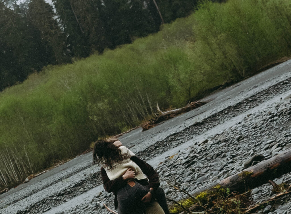 A couple embraces on the grey rocky riverbank of the Hoh River, Olympic National Park, her dark curly hair and cream knit sweater catching the damp air as he pulls her close. Shot from above at a diagonal, the wet stone riverbed stretches behind them alongside a vivid strip of bright green riverside willows and dark conifers in the distance.