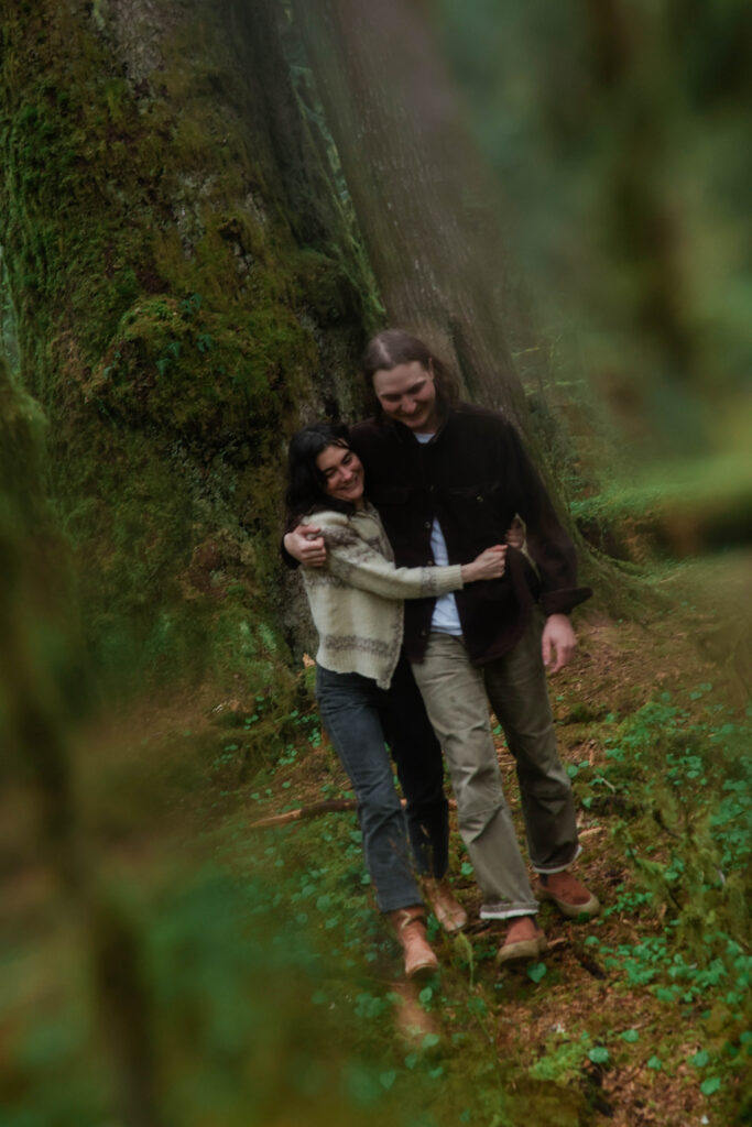 A couple laughs and embraces against the base of a massive moss-covered old-growth tree in the Hoh Rainforest, Olympic National Park. She is wearing a cream knit sweater and jeans, he is in a dark jacket and olive trousers, both in sturdy boots. The shot is framed through soft out-of-focus ferns and foliage in the foreground.