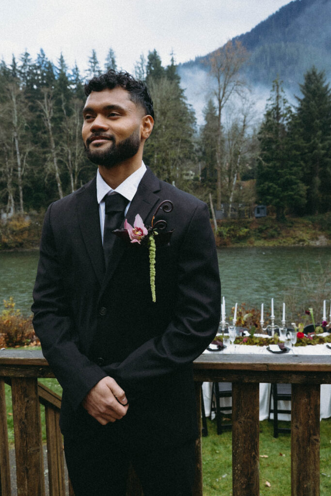 groom in dark suit with orchid boutonniere standing in front of misty mountain river Washington State elopement