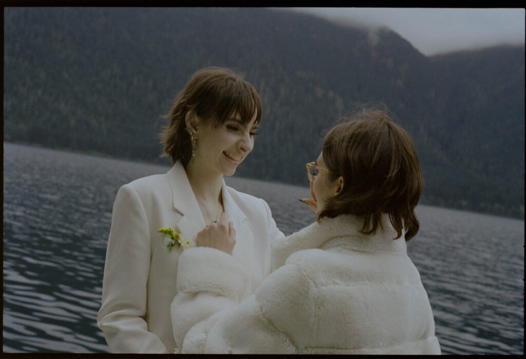 Two brides sharing a quiet moment on the shore of Lake Crescent during their Olympic National Park elopement, one in a white blazer with yellow wildflower boutonniere and one in a white faux fur coat, with the deep blue lake and forested Olympic ridgeline behind them.

