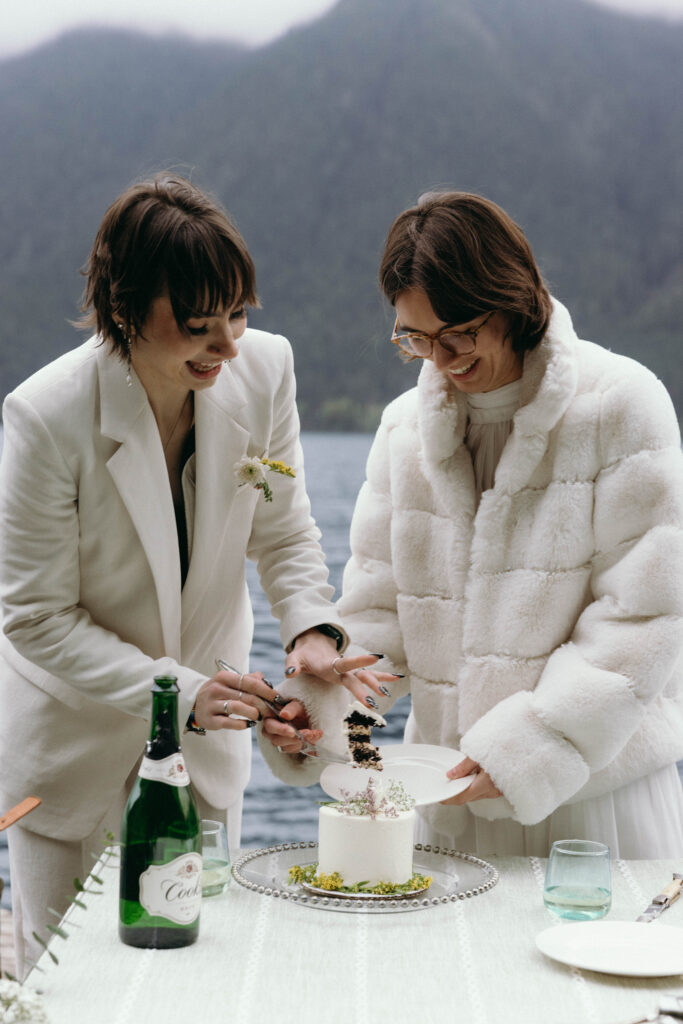 Two brides cutting their wedding cake on the shore of Lake Crescent during their Olympic National Park elopement, one in a white blazer and one in a white faux fur coat, with a single-tier white cake decorated with wildflowers, champagne bottle, and the misty Lake Crescent ridgeline visible in the background.