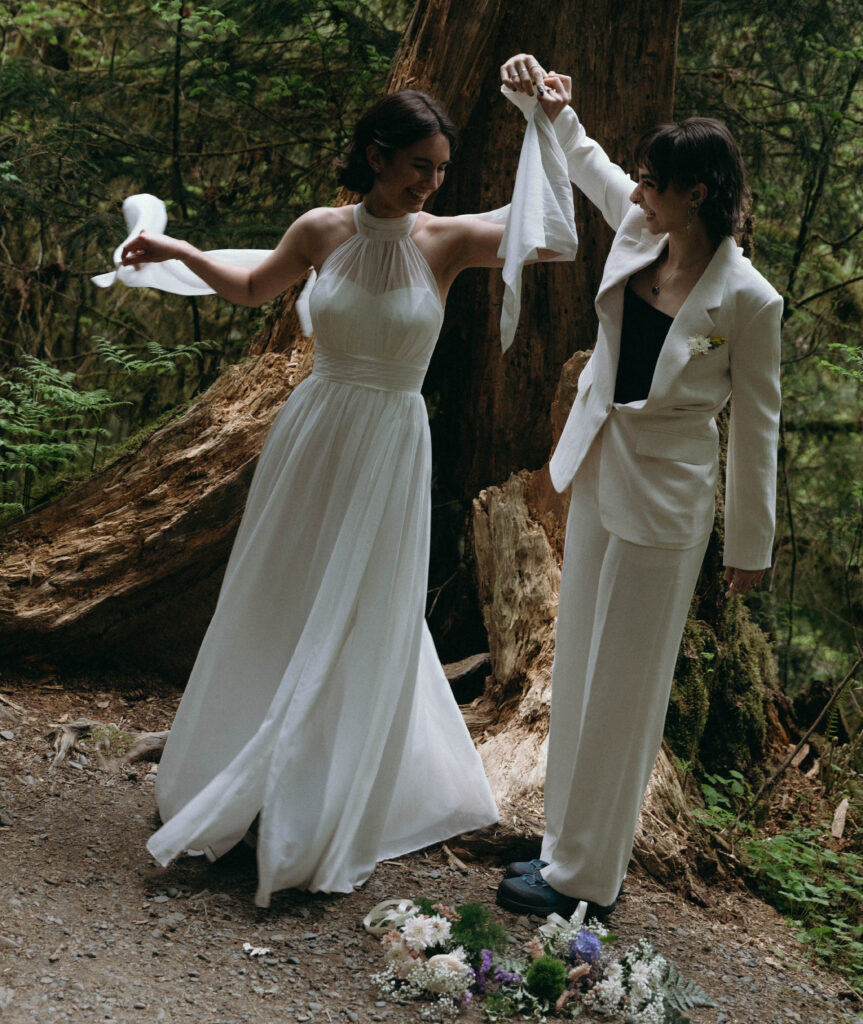 Two brides laughing and spinning together in front of a fallen old-growth cedar during their Olympic National Park elopement, one in a flowing white halter gown and one in a white suit with boutonniere, wildflower bouquets on the ground at their feet on the Hoh Rainforest trail.
