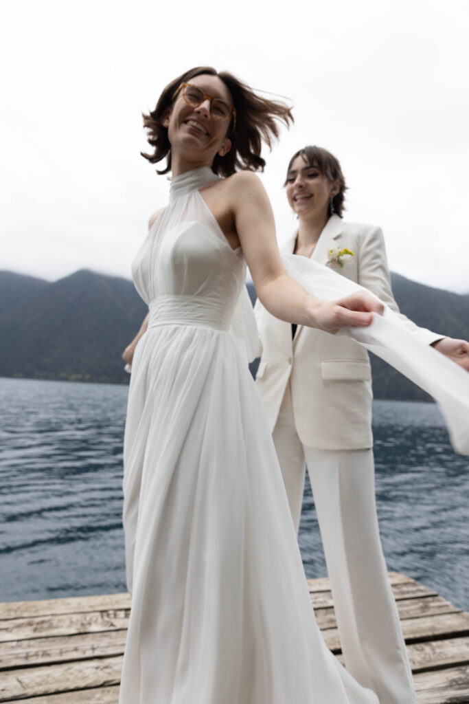 Low-angle shot of two brides laughing on the Lake Crescent dock during their Olympic National Park elopement, hair and wedding gown caught in the wind, with the steel-blue lake and Olympic mountain ridgeline behind them.


