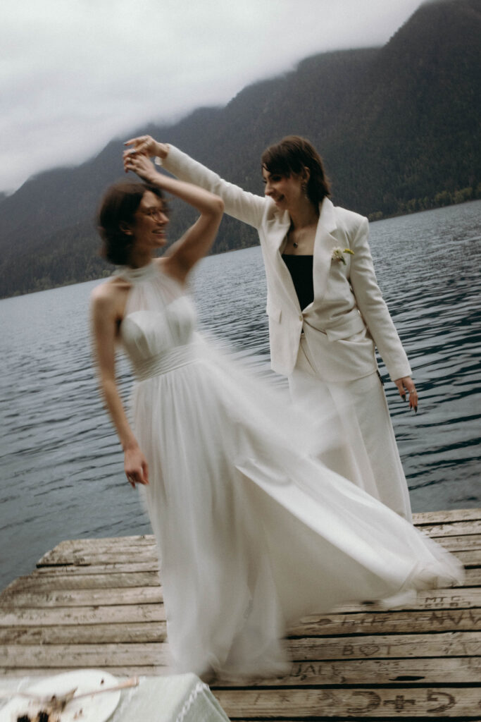 Two brides dancing and spinning on a weathered dock at Lake Crescent during their Olympic National Park elopement, wedding gown flowing in motion blur, with the deep blue-green lake and misty Olympic mountain ridgeline in the background.