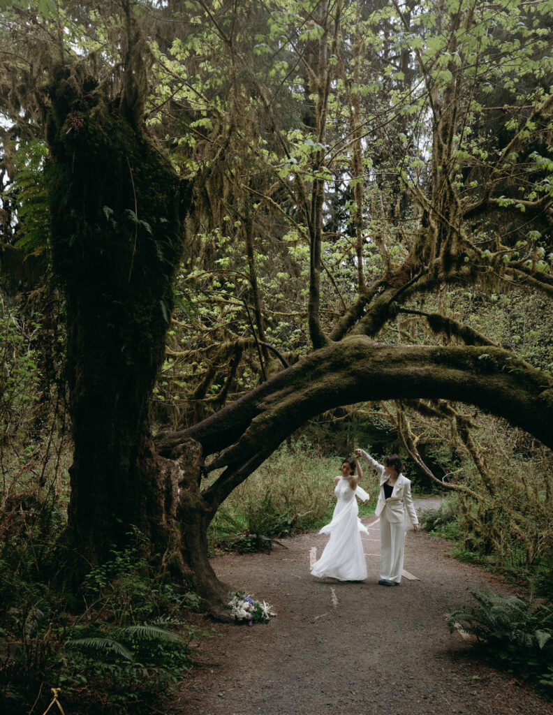 Two brides dancing under a massive moss-covered bigleaf maple arch on the Hall of Mosses trail during their Hoh Rainforest elopement in Olympic National Park, one in a flowing white halter gown and one in a white suit, with their wildflower bouquet resting on the path nearby.