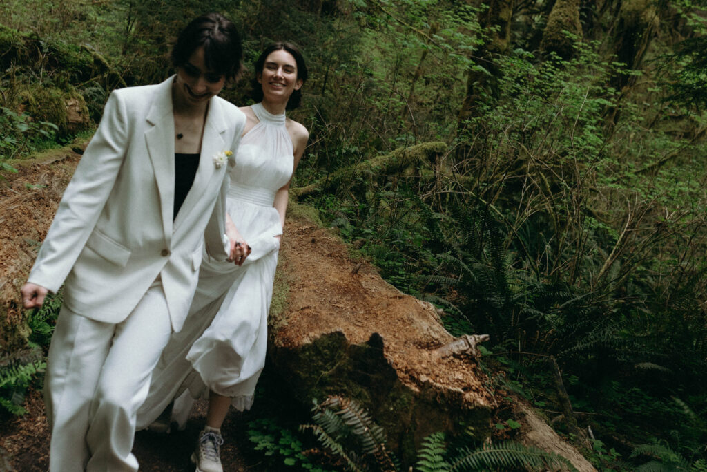 Two brides holding hands and laughing while navigating a fallen log on the Hoh Rainforest trail during their Olympic National Park elopement, surrounded by sword ferns and moss-covered old-growth trees in the Washington state temperate rainforest.


