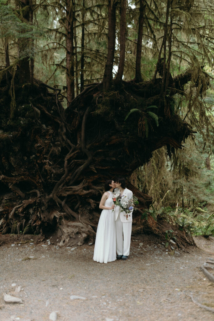 Two brides sharing a kiss in front of a massive fallen tree root system in the Hoh Rainforest at Olympic National Park, one wearing a white halter gown and the other a white suit, holding a wildflower bouquet with moss-covered trees in the background.