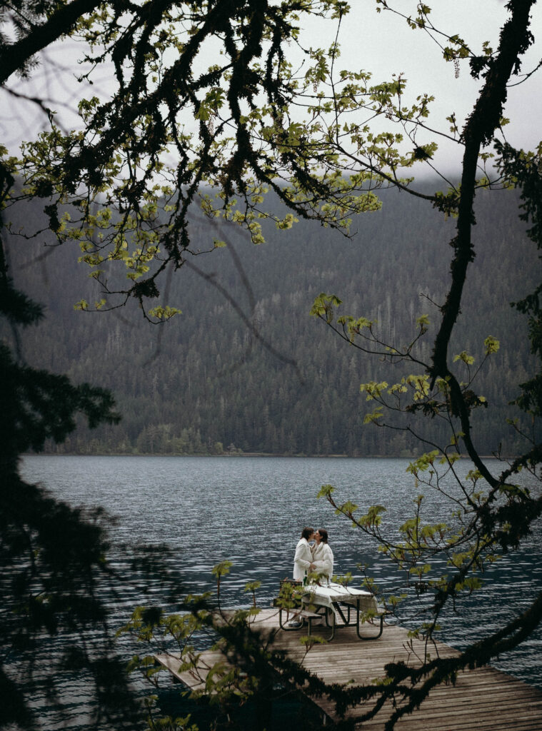 Wide shot of two brides kissing at a picnic table on the Lake Crescent dock during their Olympic National Park elopement, framed by budding bigleaf maple branches in the foreground with the blue-green lake and densely forested Olympic ridgeline stretching behind them.