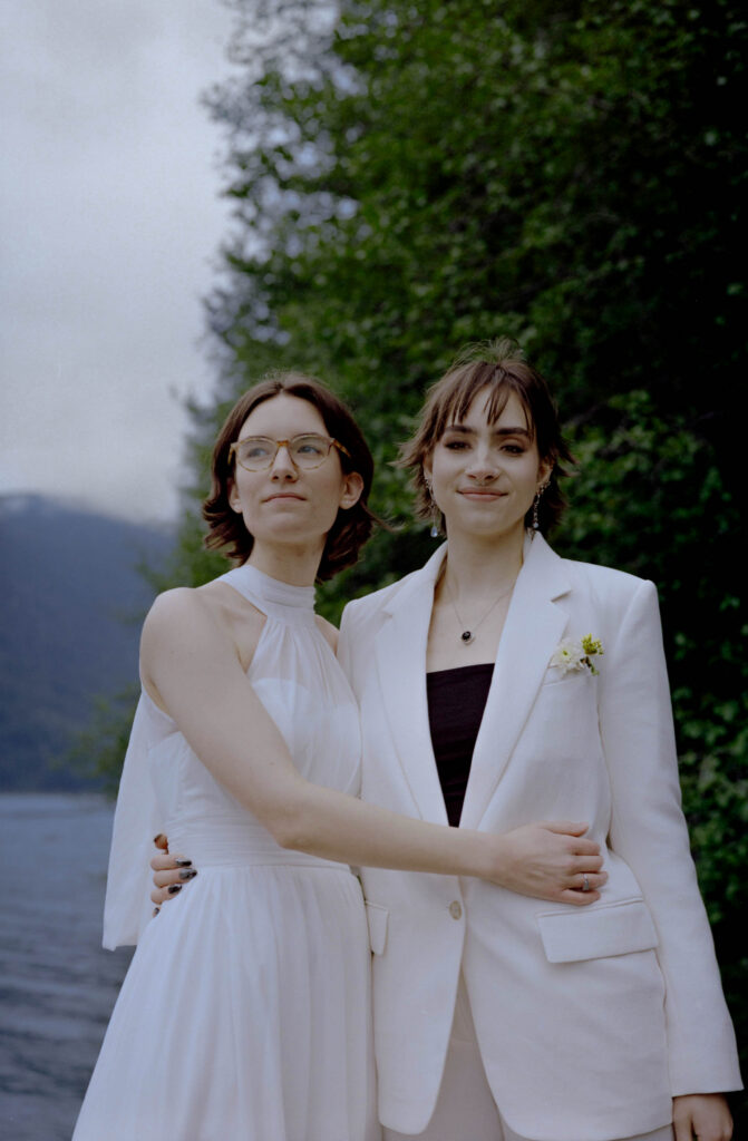 Portrait of two brides on the shore of Lake Crescent during their Olympic National Park elopement, one in a white halter gown and glasses and one in a white blazer with wildflower boutonniere, arms around each other with the lake and Olympic mountain ridgeline behind them.


