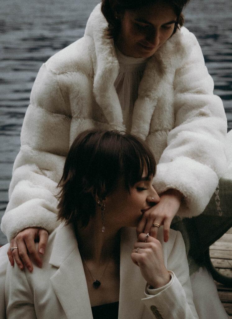 Bride in a white blazer kissing her partner's hand on the Lake Crescent dock during their Olympic National Park elopement, both wearing wedding bands, her partner seated above her in a white faux fur coat with the lake visible behind them.