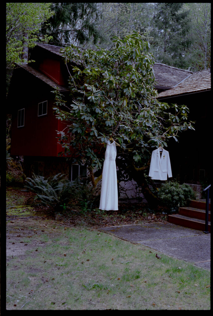 A white halter wedding gown and white suit jacket hanging from a rhododendron tree outside the Misty Valley Inn near the Hoh Rainforest, with the inn's red cedar exterior and Pacific Northwest forest visible in the background.