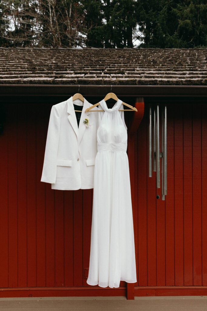 White blazer with a small floral boutonniere and white halter wedding gown hanging on wooden hangers against the red cedar exterior of the Misty Valley Inn, with a moss-covered roof and Pacific Northwest evergreens visible above.