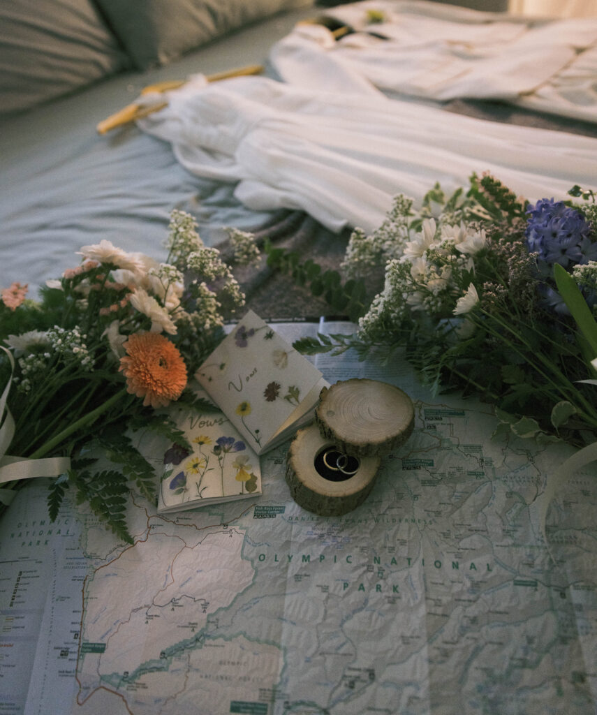 Elopement details flat lay on an Olympic National Park map, including two wildflower bouquets, pressed flower vow books, wedding bands in a wood slice ring box, and a white wedding gown in the background, styled on a bed at the Misty Valley Inn.

