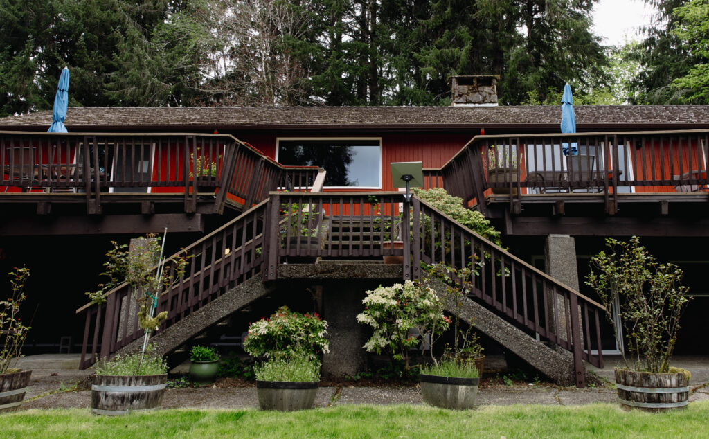 Exterior of the Misty Valley Inn near the Hoh Rainforest, showing the red cedar building with dual staircases, wood decks with blue umbrellas, potted flowering plants, and tall Pacific Northwest evergreens in the background.