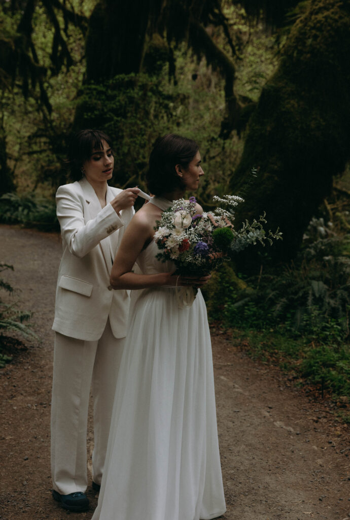Bride in a white suit fastening the halter tie of her partner's wedding gown on the Hall of Mosses trail during their Hoh Rainforest elopement, with a lush wildflower bouquet and moss-covered bigleaf maples in the background.